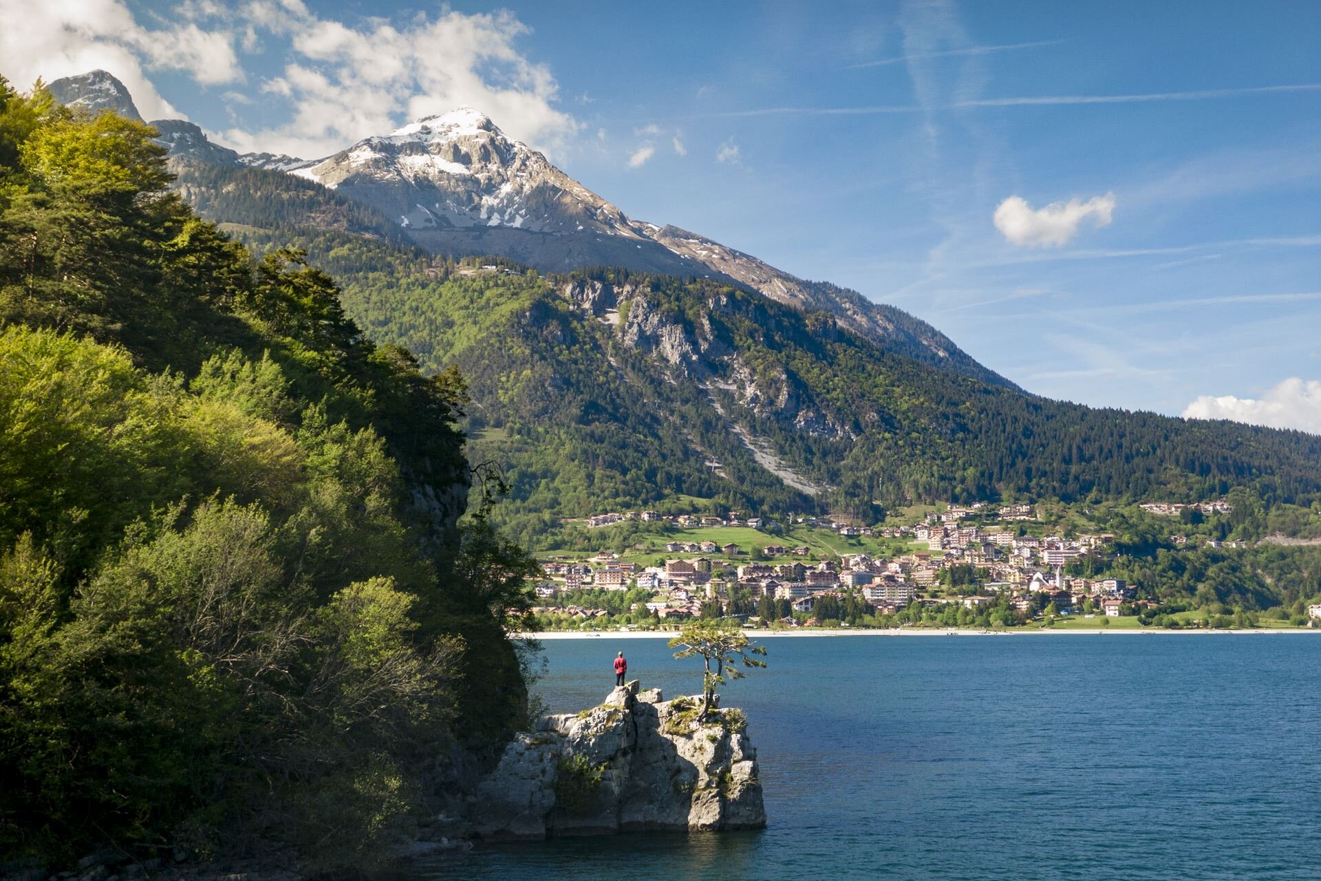 A person in a red jacket stands on a small, rocky promontory overlooking the blue waters of Lake Molveno in spring. In the background, the town of Molveno is nestled at the foot of an imposing mountain with a snow-capped peak, under a blue sky. | © Filippo Frizzera, 2019