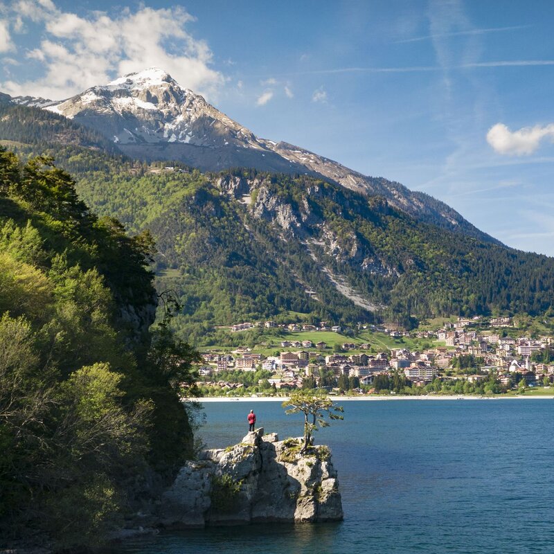 Una persona con una giacca rossa sta in piedi su un piccolo promontorio roccioso che si affaccia sulle acque blu del Lago di Molveno in primavera. Sullo sfondo, il paese di Molveno è adagiato ai piedi di un'imponente montagna con la cima innevata, sotto un cielo azzurro. | © Filippo Frizzera, 2019