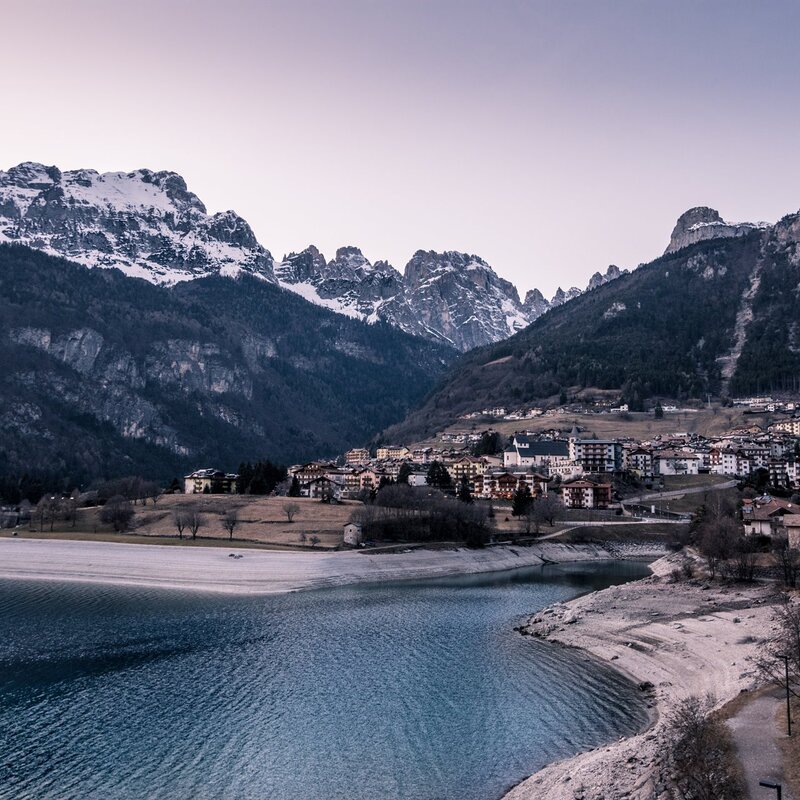 Ein ruhiger, tiefblauer See nimmt den unteren Teil des Bildes ein, umgeben von einem Strand aus hellen Kieselsteinen. Im Hintergrund liegt Molveno: Häuser mit dunklen Dächern, niedrige Gebäude und eine sichtbare Kirche mit einem Glockenturm. Hinter dem Dorf erheben sich imposant die Brenta-Dolomiten, bedeckt von dunklen Wäldern und einigen schneebedeckten Gipfeln. Der Himmel ist klar und verfärbt sich in helle Töne. Bei Sonnenuntergang ist die Atmosphäre ruhig und kalt, typisch für einen Wintertag. | © Benedetta Bernardeschi, 2020
