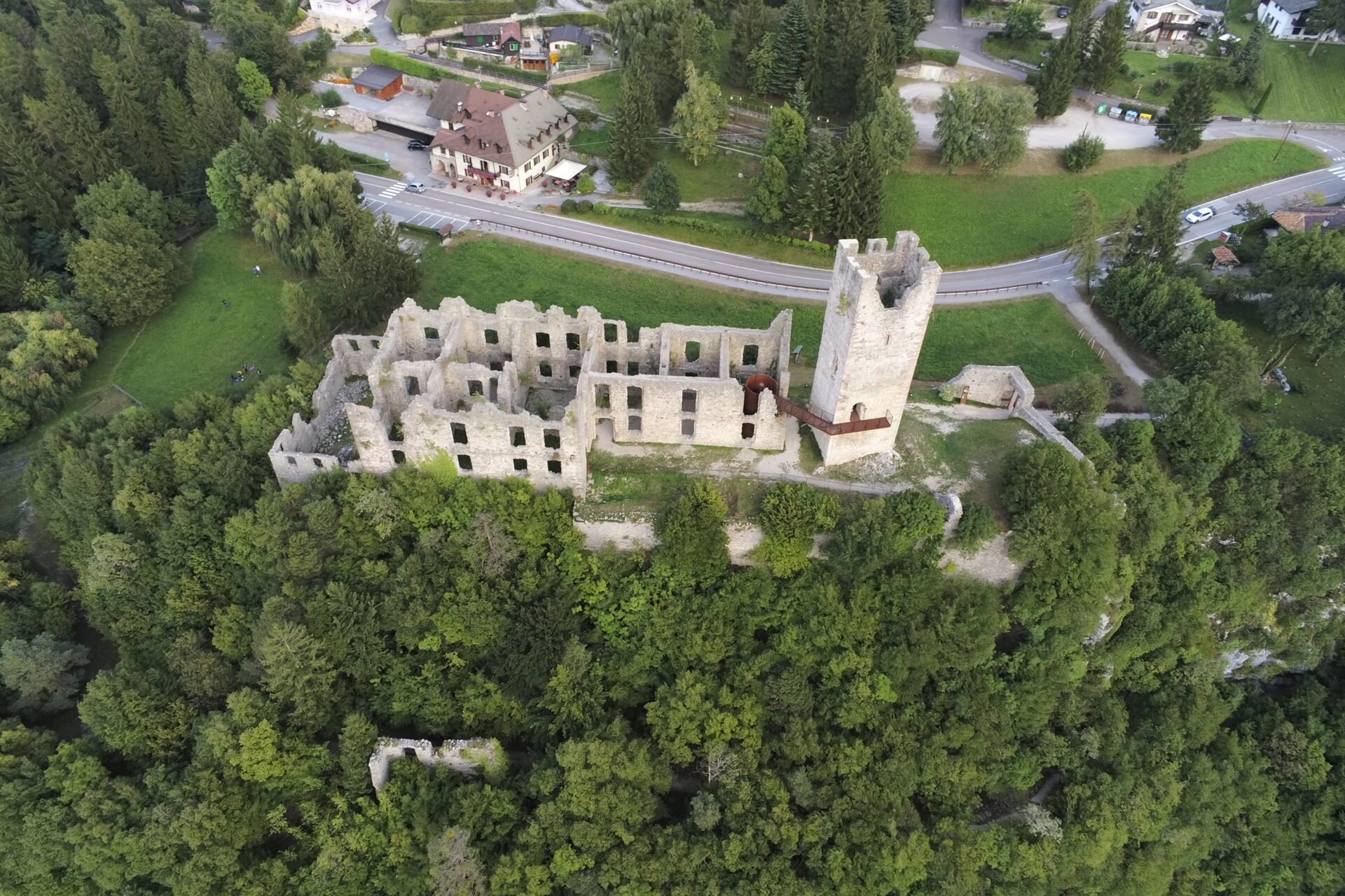 An ancient ruined castle stands atop a verdant hill, with trees and grass growing between its crumbling walls. A paved road meanders at the foot of the hill, passing a few houses scattered around the surrounding landscape. | © Alessio Mattei, 2019