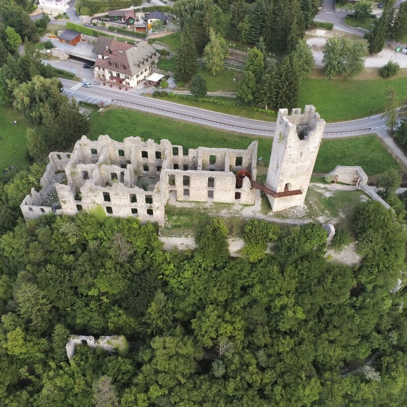 Un antico castello in rovina si erge in cima a una collina verdeggiante, con alberi e l'erba che crescono tra le sue mura fatiscenti. Una strada asfaltata serpeggia ai piedi della collina, passando vicino ad alcune case sparse nel paesaggio circostante. | © Alessio Mattei, 2019