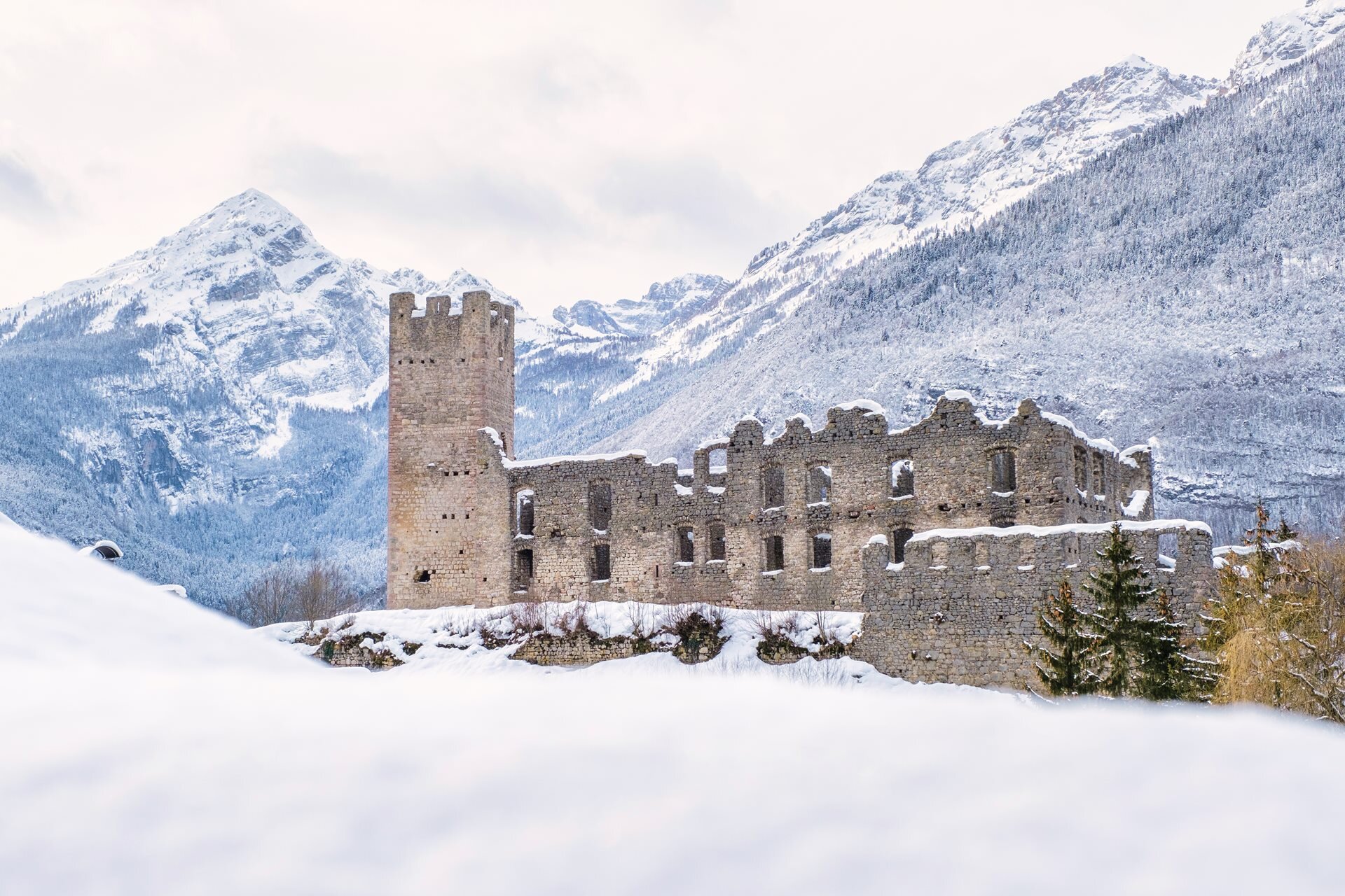 Die schneebedeckten Ruinen einer alten Burg heben sich von den schneebedeckten Bergen der Brenta-Dolomiten ab. Die teilweise eingestürzten Mauern und Türme der Burg ragen aus dem Schnee, stumme Zeugen der Vergangenheit. | © Filippo Frizzera, 2021