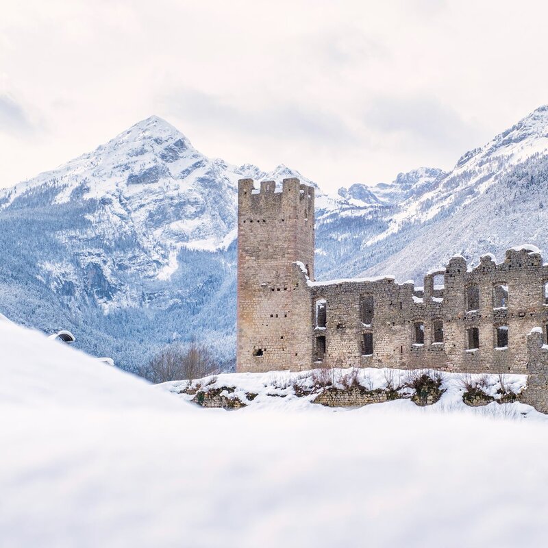 Le rovine innevate di un antico castello si stagliano contro uno sfondo di montagne innevate, le Dolomiti di Brenta. Le mura e le torri del castello, parzialmente crollate, emergono dalla neve, testimoni silenziose del passato. | © Filippo Frizzera, 2021