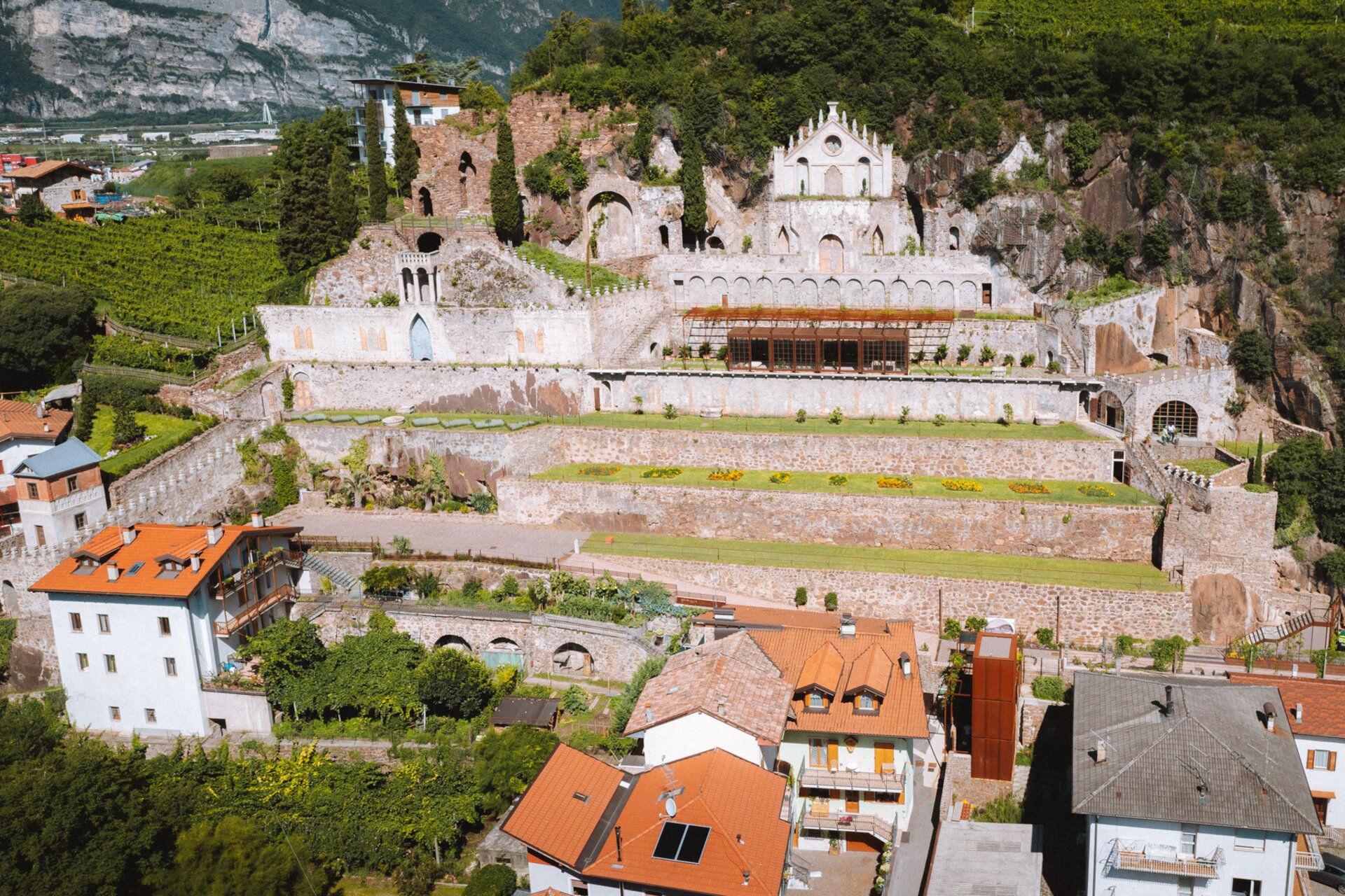 Un antico complesso, con mura in pietra e una facciata decorata con archi e un rosone, domina un pendio terrazzato con giardini curati.  Ai piedi della collina, case con tetti rossi si affacciano sul complesso, creando un contrasto tra antico e moderno. | © Nicola Cagol, 2021