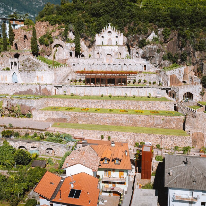 Ein antiker Komplex mit Steinmauern und einer mit Bögen und einer Rosette verzierten Fassade dominiert einen terrassenförmigen Hang mit gepflegten Gärten.  Am Fuße des Hügels überragen Häuser mit roten Dächern den Komplex und bilden einen Kontrast zwischen Antike und Moderne. | © Nicola Cagol, 2021