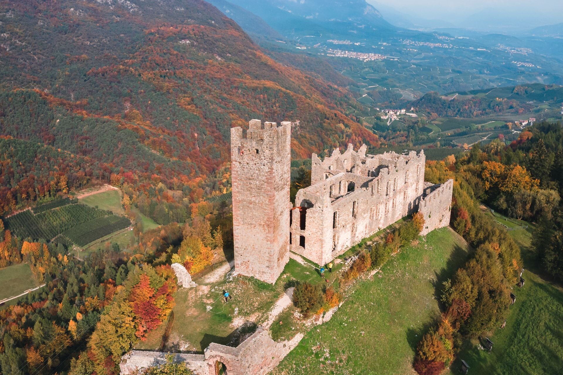 Vista aerea di Castel Belfort, un'antica rovina medievale immersa nel foliage autunnale delle montagne trentine. La torre e le mura di pietra contrastano con i colori caldi della vegetazione, mentre la valle sottostante si estende tra campi coltivati e piccoli borghi. | © Enrico Baroni, 2020