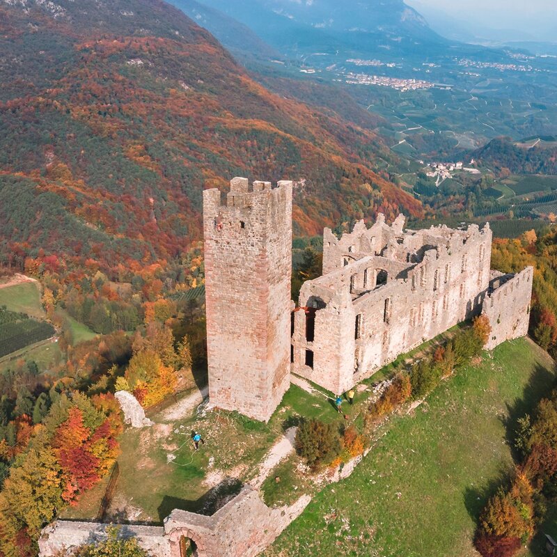 Aerial view of Castel Belfort, an ancient medieval ruin nestled in the autumn foliage of the mountains of Trentino. The tower and stone walls contrast with the warm colors of the vegetation, while the valley below stretches between cultivated fields and small villages. | © Enrico Baroni, 2020