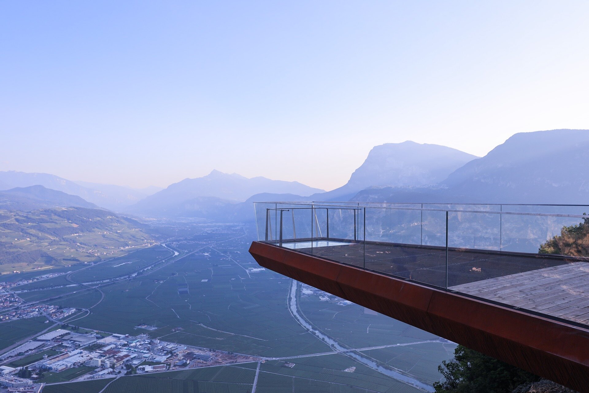 The Skywalk of Monte di Mezzocorona, a panoramic platform suspended in the air with a glass railing. From the viewpoint, there is a vast aerial view over the Piana Rotaliana, with its cultivated fields, towns, and distant mountains. | © @worldmappers, 2023