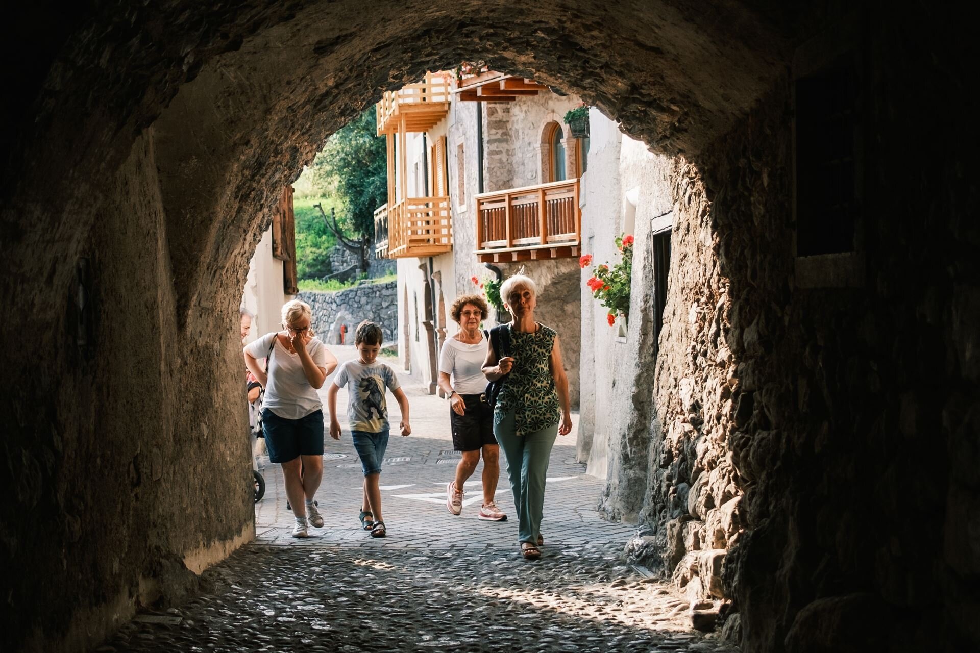 Eine Gruppe von Personen, darunter eine junge Person, tritt aus einer engen, dunklen Steingasse auf eine sonnige, kopfsteingepflasterte Straße. Im Hintergrund sind die Fassaden alter Häuser mit Holzbalkonen zu sehen. | © Filippo Frizzera, 2024