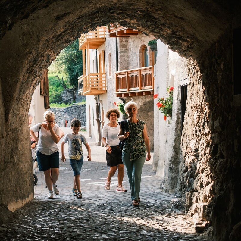 Eine Gruppe von Personen, darunter eine junge Person, tritt aus einer engen, dunklen Steingasse auf eine sonnige, kopfsteingepflasterte Straße. Im Hintergrund sind die Fassaden alter Häuser mit Holzbalkonen zu sehen. | © Filippo Frizzera, 2024
