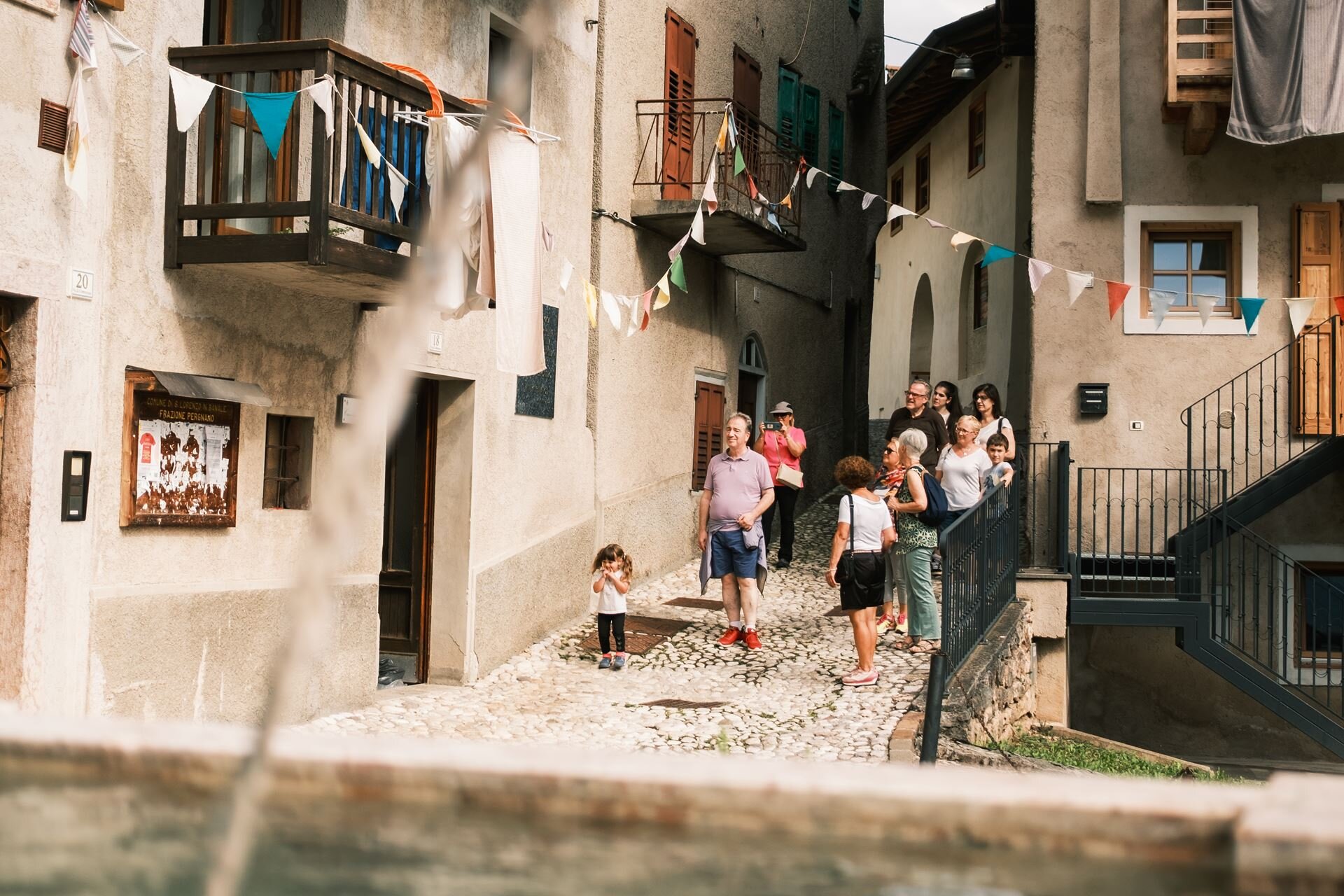 Eine Gruppe von Personen steht in einer engen Gasse eines alten Dorfes, das von Gebäuden mit rauhem Putz und bunten Girlanden geprägt ist. | © Filippo Frizzera, 2024