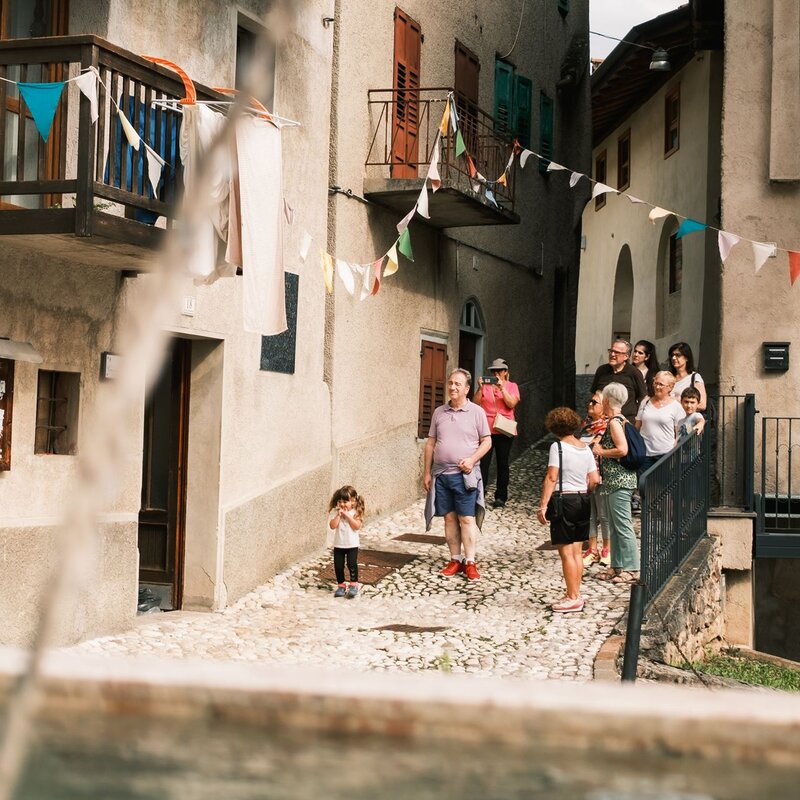Un gruppo di persone sosta in una stretta via di un borgo antico, caratterizzata da edifici con intonaco grezzo e festoni colorati. | © Filippo Frizzera, 2024
