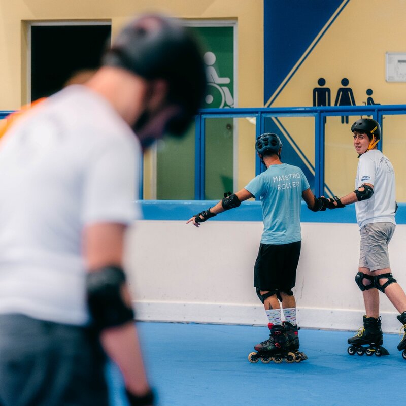 All'interno di una pista da pattinaggio dell'Andalo Life, un istruttore con la maglia azzurra "Maestro Roller" guida un ragazzo sui rollerblade tenendolo per mano. Entrambi indossano casco e protezioni. In primo piano, altre persone sfuocate partecipano all'attività. | © Mirko Perli, 2025