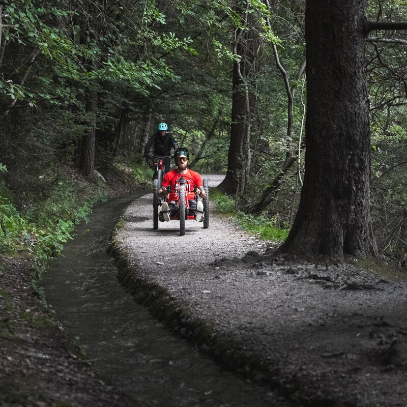 Un uomo con una maglietta rossa e occhiali da sole guida una handbike a tre ruote lungo un sentiero sterrato pianeggiante che attraversa un bosco ombroso. Poco dietro di lui si nota un secondo ciclista in sella a una mountain bike. Sul lato sinistro del tracciato scorre un piccolo canale d'acqua artificiale delimitato da sponde in pietra. | © Camilla Pizzini, 2024