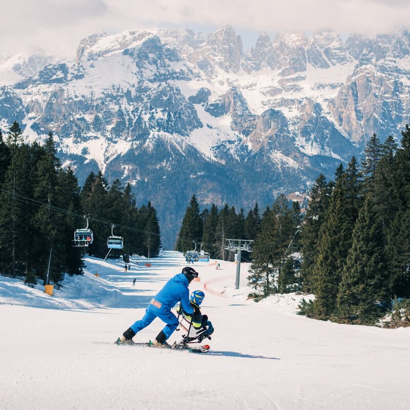 Veduta ampia di una pista da sci. Al centro, un maestro in azzurro guida un sit-ski in discesa. A sinistra scorre una seggiovia sopra i pini. Sullo sfondo si innalzano imponenti vette rocciose frastagliate. | © Filippo Frizzera, 2026