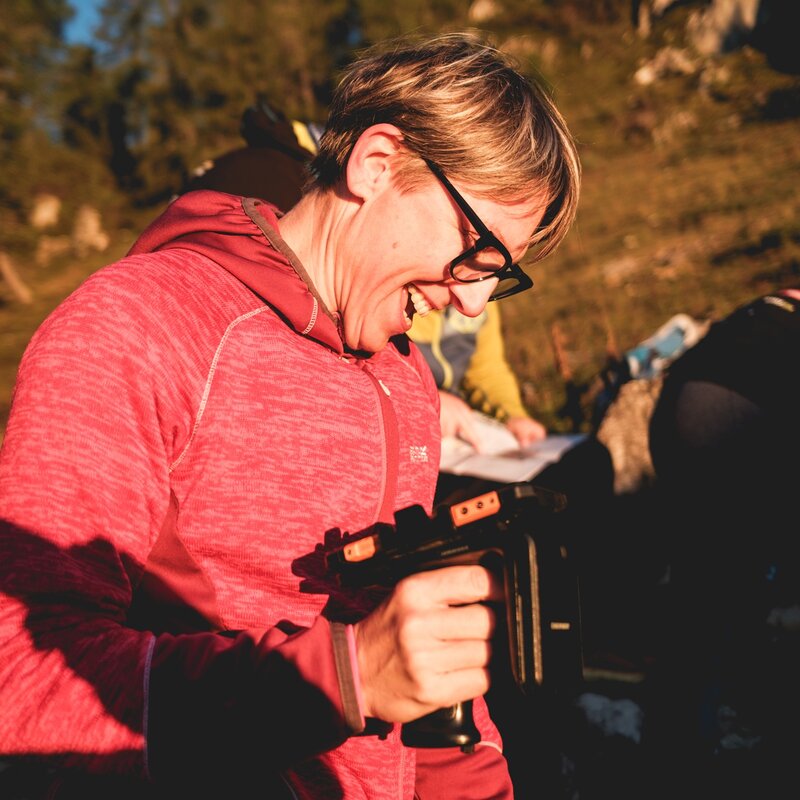 Una volontaria con occhiali e felpa tecnica fucsia sorride con entusiasmo durante il Dolomiti Brenta Trail. Tiene in mano un dispositivo elettronico mentre la luce calda del tramonto illumina la scena in montagna. | © Filippo Frizzera, 2025