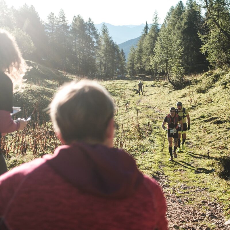 Due trail runner avanzano lungo un sentiero erboso in salita, seguiti dallo sguardo dei volontari in primo piano. La scena è immersa nella luce calda del mattino tra i boschi della Paganella. | © Filippo Frizzera, 2025
