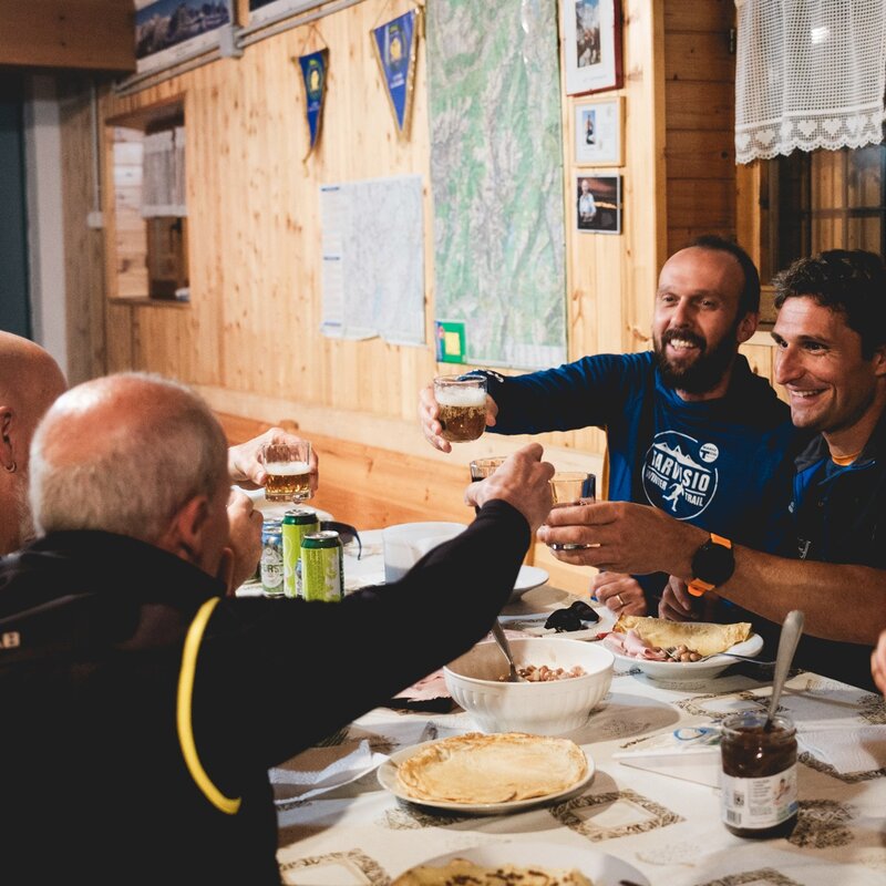 Un gruppo di uomini siede attorno a una tavola imbandita all'interno di un rifugio in legno. Sorridono e brindano con dei bicchieri in un'atmosfera calda e amichevole la sera prima dell'evento sportivo. | © Filippo Frizzera, 2025
