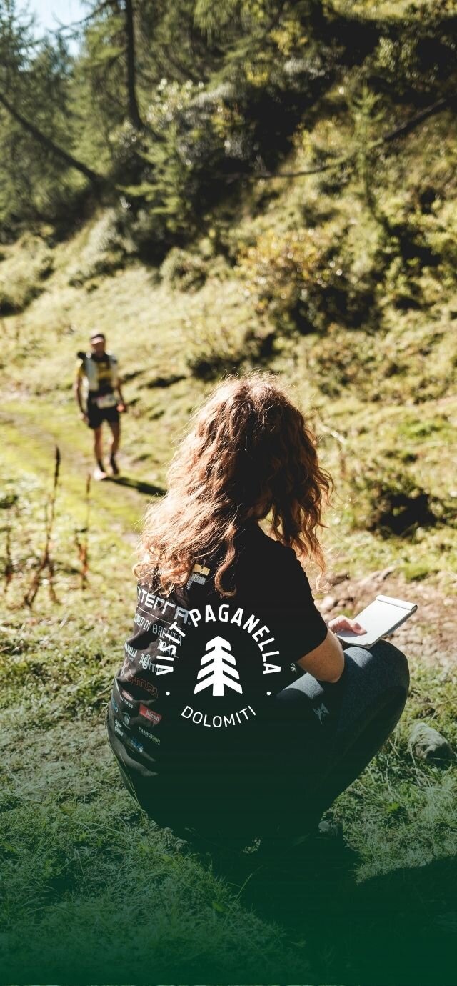 Inquadratura verticale di una volontaria di spalle, mentre prende appunti su un block notes. Sullo sfondo, un atleta percorre il sentiero nel bosco illuminato dal sole.  Logo "Visit Paganella Dolomiti" in primo piano. | © Filippo Frizzera, 2025