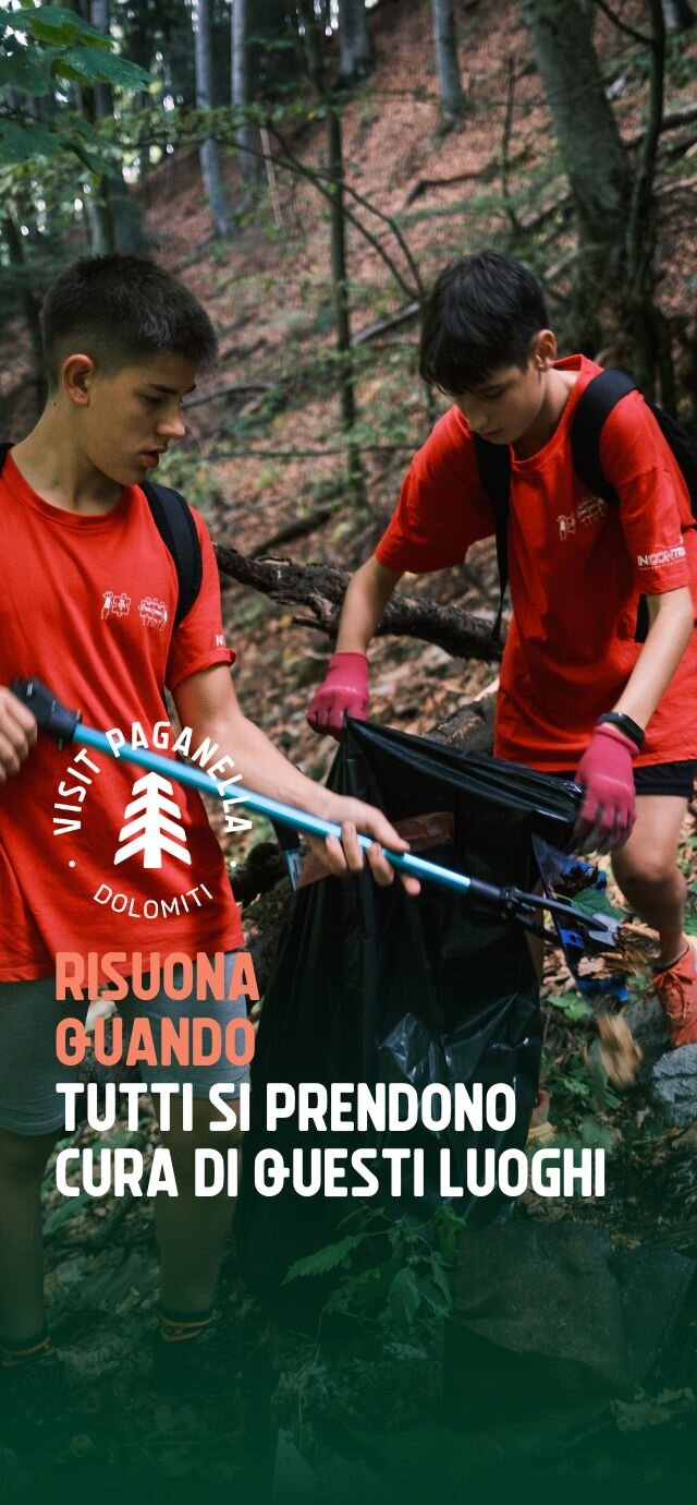 Due giovani volontari in maglietta rossa raccolgono rifiuti nel bosco della Paganella con pinze e sacchi neri, prendendosi cura del territorio. Sull'immagine c'è il logo della destinazione e una frase che recita 'RISUONA QUANDO TUTTI SI PRENDONO CURA DI QUESTI LUOGHI'. | © Filippo Frizzera, 2024