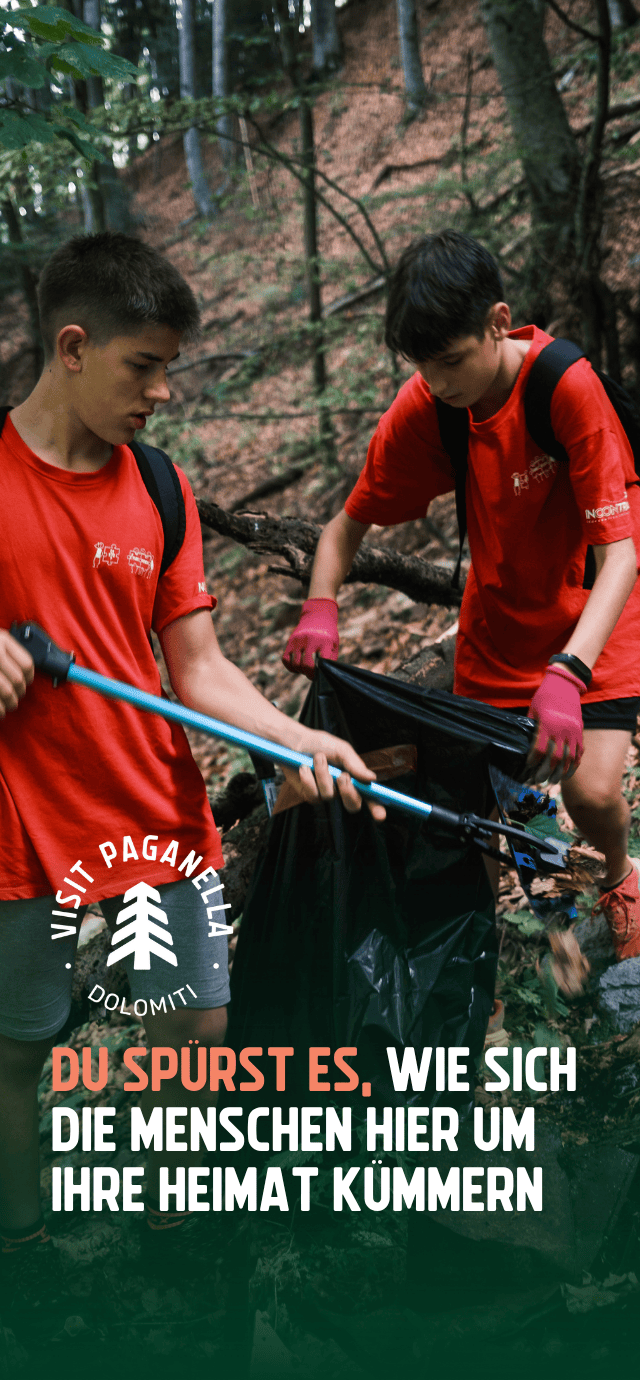 Zwei junge Menschen in roten T-Shirts und mit Rucksäcken sind in einem Wald mit Aufräumarbeiten beschäftigt. Eine Person hebt mit einer Teleskopzange etwas vom Boden auf, während die andere einen großen schwarzen Müllsack offen hält. Über dem Bild sind ein Logo und der Schriftzug „DU SPÜRST ES, WIE SICH DIE MENSCHEN HIER UM IHRE HEIMAT KÜMMERN” zu sehen. | © Filippo Frizzera, 2024