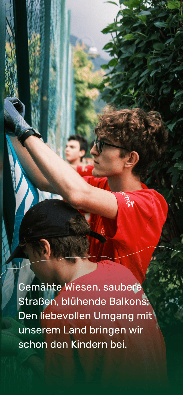 Einige junge Leute in roten T-Shirts und Arbeitshandschuhen sind damit beschäftigt, eine Plane oder ein Banner an einem Metallzaun im Freien anzubringen. Im Vordergrund konzentriert sich eine Person mit Brille auf ihre Arbeit, während im Vordergrund ein Text zu lesen ist: „Gemähte Wiesen, saubere Straßen, blühende Balkons: Den liebevollen Umgang mit unserem Land bringen wir schon den Kindern bei.“ | © Filippo Frizzera, 2024