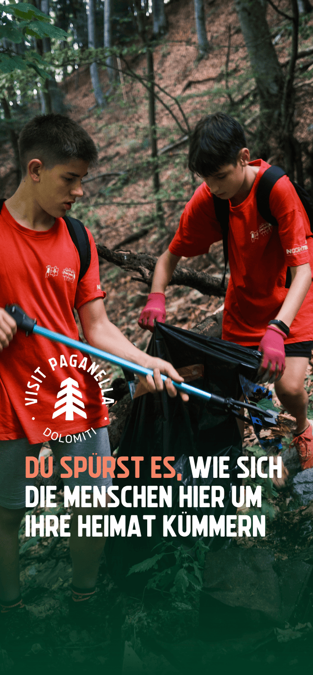 Zwei junge Menschen in roten T-Shirts und mit Rucksäcken sind in einem Wald mit Aufräumarbeiten beschäftigt. Eine Person hebt mit einer Teleskopzange etwas vom Boden auf, während die andere einen großen schwarzen Müllsack offen hält. Über dem Bild sind ein Logo und der Schriftzug „DU SPÜRST ES, WIE SICH DIE MENSCHEN HIER UM IHRE HEIMAT KÜMMERN” zu sehen. | © Filippo Frizzera, 2024