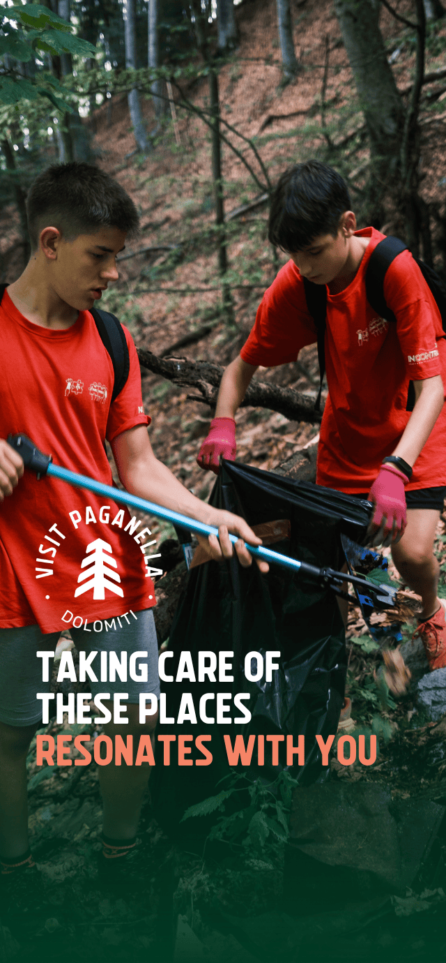Two young people, wearing red T-shirts and backpacks, are engaged in a clean-up activity in a forest. One person uses telescopic tongs to pick something up from the ground, while the other holds open a large black rubbish bag. Overlaying the image are a logo and the words “TAKING CARE OF THESE PLACES RESONATES WITH YOU”. | © Filippo Frizzera, 2024