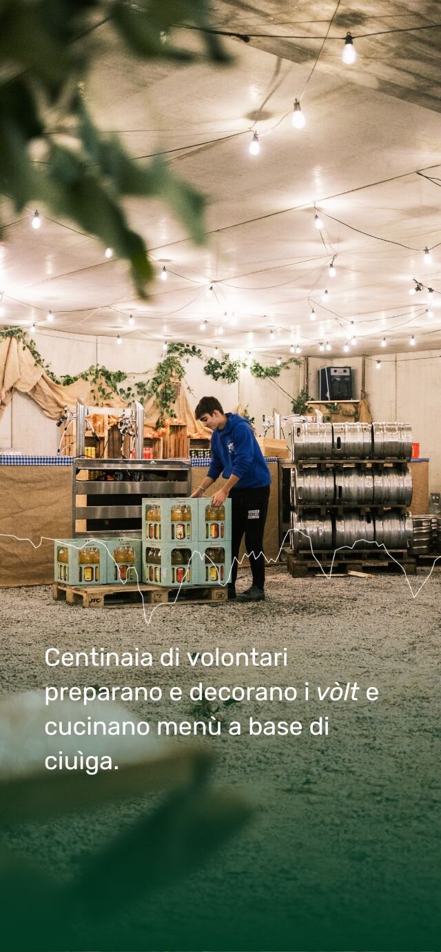 Un giovane volontario allestisce uno stand gastronomico all'interno di un tendone per la Sagra della Ciuìga. Attorno a lui, fusti di birra e un bancone bar, sotto le luci di una festa.  Testo in sovrimpressione: Centinaia di volontari preparano e decorano i vòlt e cucinano menù a base di ciuìga. | © Filippo Frizzera