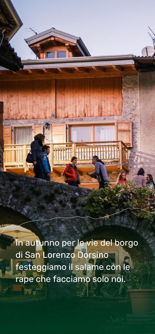 Un gruppo di persone si gode l'atmosfera della Sagra della Ciuìga su un antico ponte in pietra, nelle vie del borgo di San Lorenzo Dorsino. Sullo sfondo, una tipica casa alpina in legno e sasso.  Testo in sovrimpressione: In autunno per le vie del borgo di San Lorenzo Dorsino festeggiamo il salame con le rape che facciamo solo noi. | © Filippo Frizzera, 2024
