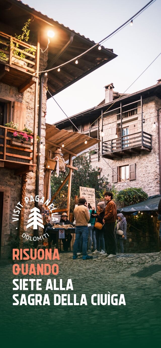 Persone riunite in una piazzetta lastricata di San Lorenzo Dorsino, illuminate dalle luci di un filo appeso tra le case in pietra del borgo. La scena cattura un momento conviviale attorno a uno stand della Sagra della Ciuìga.  Testo in sovrimpressione: Il logo "VISIT PAGANELLA " e la scritta "RISUONA QUANDO SIETE ALLA SAGRA DELLA CIUÌGA". | © Filippo Frizzera, 2024