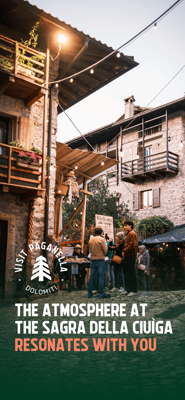 People gather around a food stall in a historic village square during the Sagra della Ciuìga festival in San Lorenzo Dorsino. The scene is lit by string lights hung between the stone houses.  Overlay Text:  THE ATMOSPHERE AT THE SAGRA DELLA CIUÌGA RESONATES WITH YOU | © Filippo Frizzera, 2024