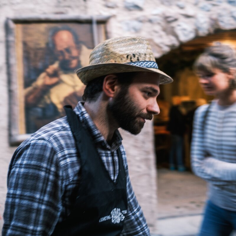 Un uomo con barba e cappello di paglia, che indossa una camicia a quadri e un gilè, cammina per le vie del borgo di San Lorenzo Dorsino. L'immagine, con un leggero effetto di mosso, cattura l'atmosfera dinamica della Sagra della Ciuìga, con altre persone e un ritratto appeso al muro sullo sfondo. | © Filippo Frizzera, 2024