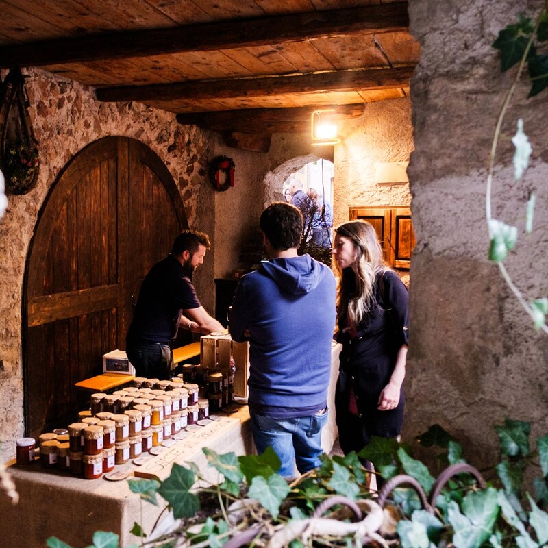 All'interno di un "vòlt" in pietra a San Lorenzo Dorsino, due clienti parlano con un venditore di prodotti tipici durante la Sagra della Ciuìga. Sul banco sono esposti numerosi vasetti di conserve, in una suggestiva cantina con travi in legno e una porta ad arco. | © Valerio Rigotti, 2022