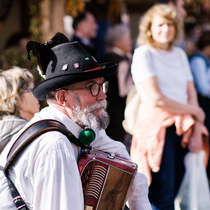 Un musicista con barba e cappello tradizionale suona la fisarmonica durante la Sagra della Ciuìga a San Lorenzo Dorsino. Il primo piano cattura l'artista in abiti folkloristici, immerso nell'atmosfera di una festa di paese con altre persone sfocate sullo sfondo. | © Valerio Rigotti, 2022