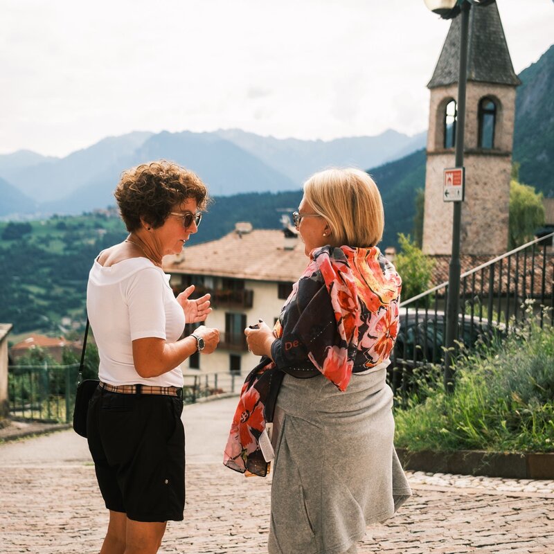 Two middle-aged women are conversing in a mountain village. One is wearing a white T-shirt, black shorts and pink trainers, with a shoulder bag and a watch on her wrist. The other is wearing a floral cover-up in orange tones, grey leggings and neon yellow trainers. In the background we see a stone bell tower with a peaked roof, houses with sloping roofs and a view of the mountains. A row of coloured flags crosses the scene, adding a festive touch. The atmosphere is relaxed and convivial. | © Filippo Frizzera, 2024