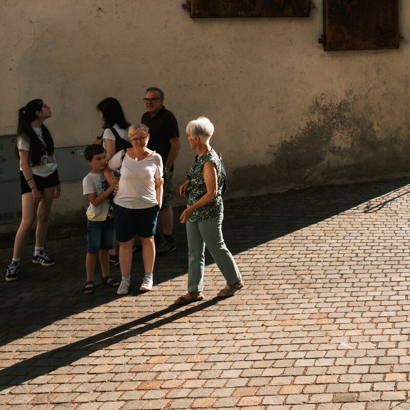 A group of six people, consisting of adults and children, is gathered in a stone-paved street, next to a clear wall on which hang three large relief works, probably made of wood, depicting historical or traditional scenes. The low sun creates long shadows on the pavement, illuminating the figures with a warm light. Two elderly women are in the foreground, one with white hair and a green suit who seems involved in a conversation, while the other, wearing a white T-shirt and glasses, is smiling. In the background, a man with glasses and two younger girls look at the wall and the artwork, while a child holds the hand of the woman in white. The atmosphere is relaxed, evoking a moment of socialisation and cultural discovery. | © Filippo Frizzera, 2024
