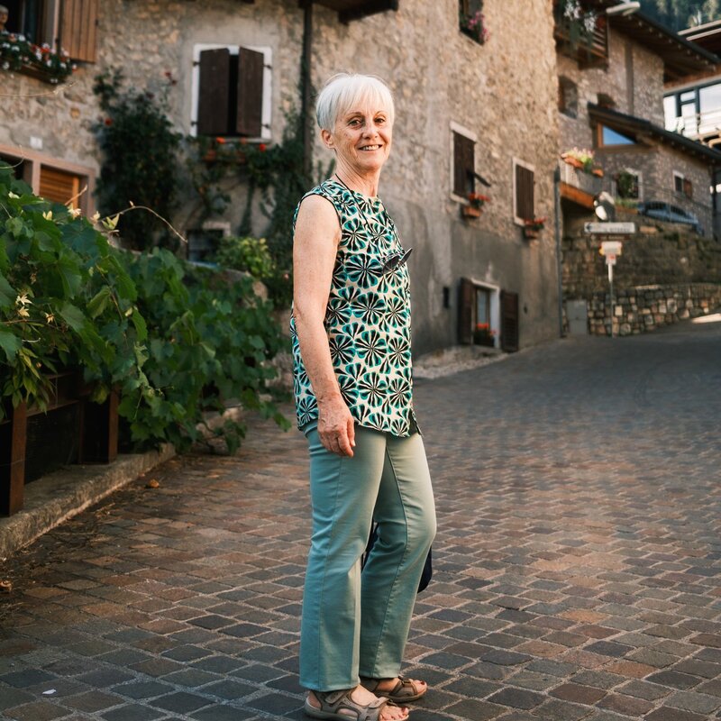 An elderly woman with short white hair smiles as she poses on a cobbled street in the village of San Lorenzo Dorsino. She wears a green outfit consisting of trousers and a sleeveless blouse with a black and white geometric pattern. On her feet she wears comfortable sandals. Behind her are stone houses with wooden windows and flower pots on the balconies. On the left, a lush vine grows along a low wall. The warm sunlight illuminates the scene, giving it a cosy and serene atmosphere. | © Filippo Frizzera, 2024