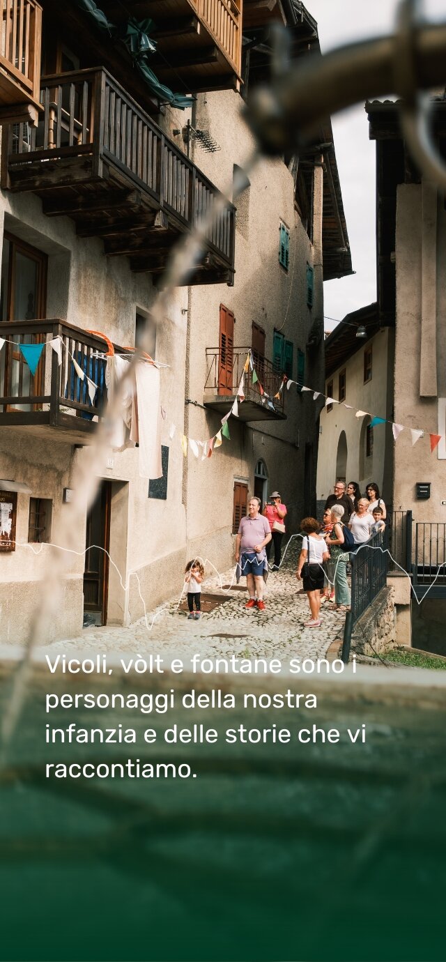 Una fontana in primo piano incornicia uno scorcio di un borgo antico. Sullo sfondo, vicoli acciottolati, case in pietra con balconi in legno e finestre colorate. Festoni di bandierine attraversano la strada. Un gruppo di persone, tra cui adulti e bambini, passeggia lungo il vicolo. Una bambina con le mani sul viso sembra giocare o proteggersi dal sole. Il testo sulla foto recita: "Vicoli, vòlti e fontane sono i personaggi della nostra infanzia e delle storie che vi raccontiamo." | © Filippo Frizzera, 2024