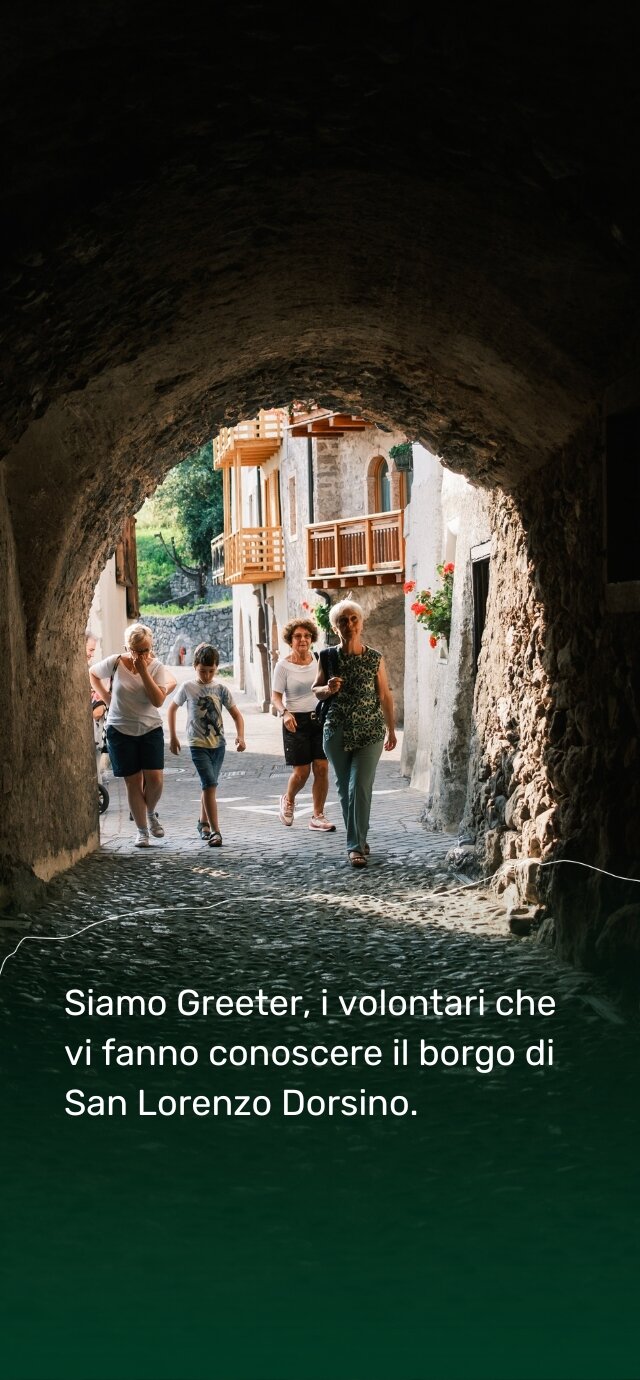 Un tunnel in pietra, con un soffitto scuro e ruvido, si apre su un vicolo soleggiato. In fondo, case di montagna con balconi in legno e fiori rossi alle finestre. Un piccolo gruppo di persone cammina verso l’uscita dell’arco, sorridendo. Una donna anziana, in primo piano, sembra guidare il gruppo. Il testo dice: "Siamo Greeter, i volontari che vi fanno conoscere il borgo di San Lorenzo Dorsino." | © Filippo Frizzera, 2024