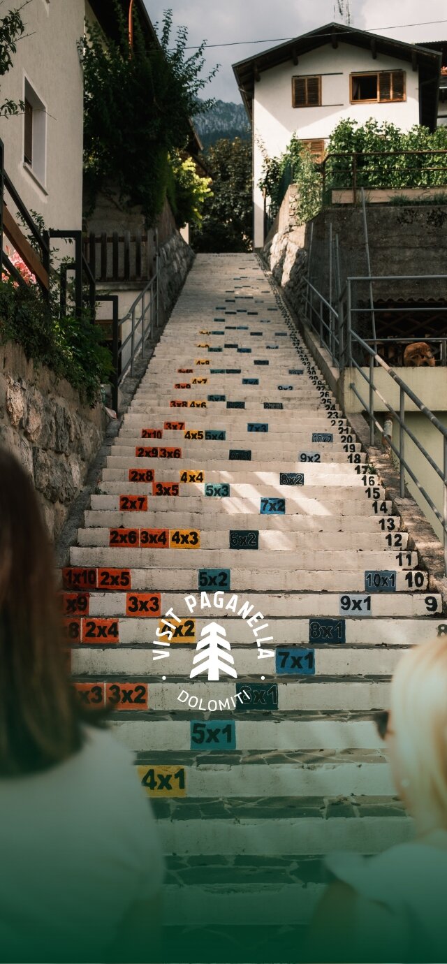 A long concrete staircase runs uphill between mountain houses. On the steps are painted colourful mathematical operations, such as multiplication tables and multiplications. Some people observe the staircase from below. The Visit Paganella logo is superimposed on the image. | © Filippo Frizzera, 2024