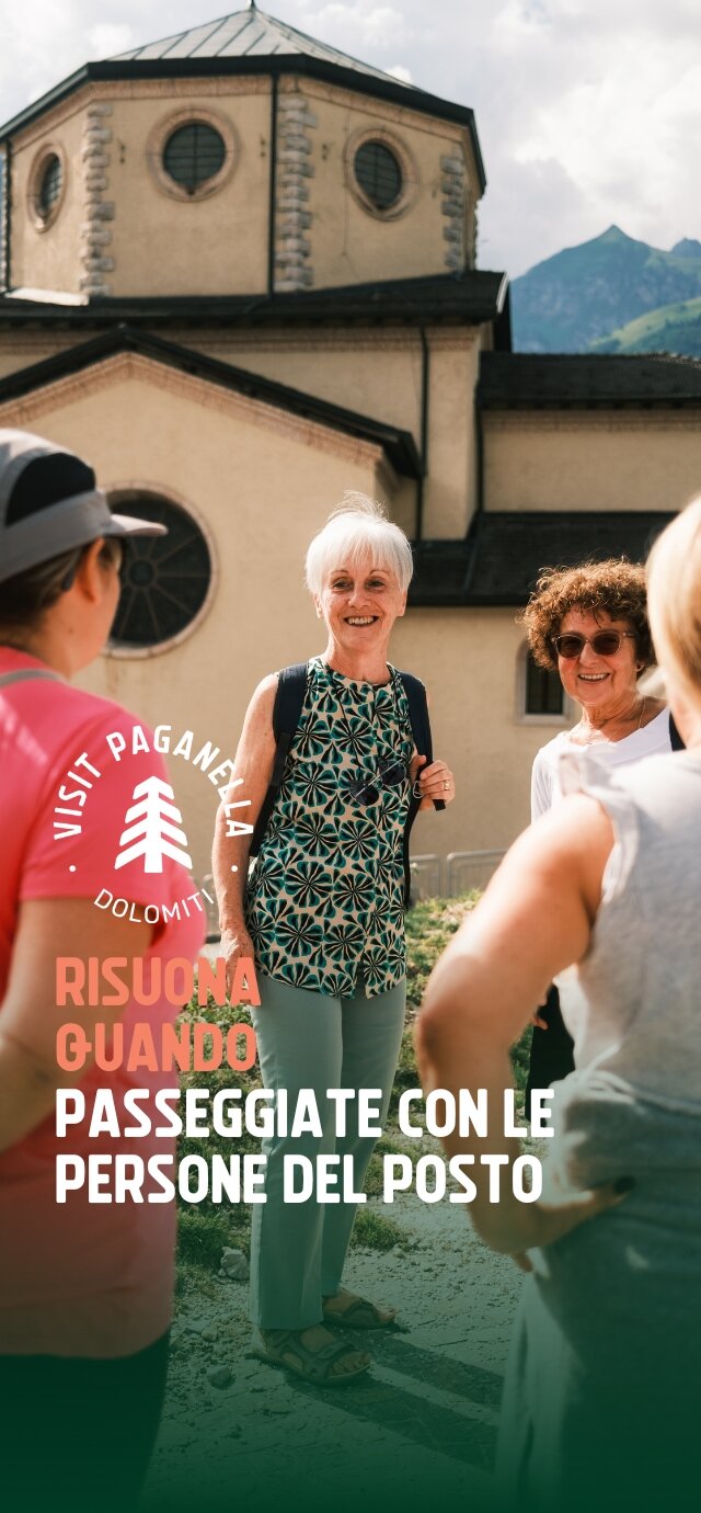 Un gruppo di persone conversa allegramente davanti a una chiesa con una grande cupola e finestre circolari. La scena è illuminata dalla luce del sole. Una donna anziana con uno zaino sorride mentre parla con altre persone. Le montagne fanno da sfondo. Il testo sulla foto recita: "Risuona quando passeggiate con le persone del posto." | © Filippo Frizzera, 2024