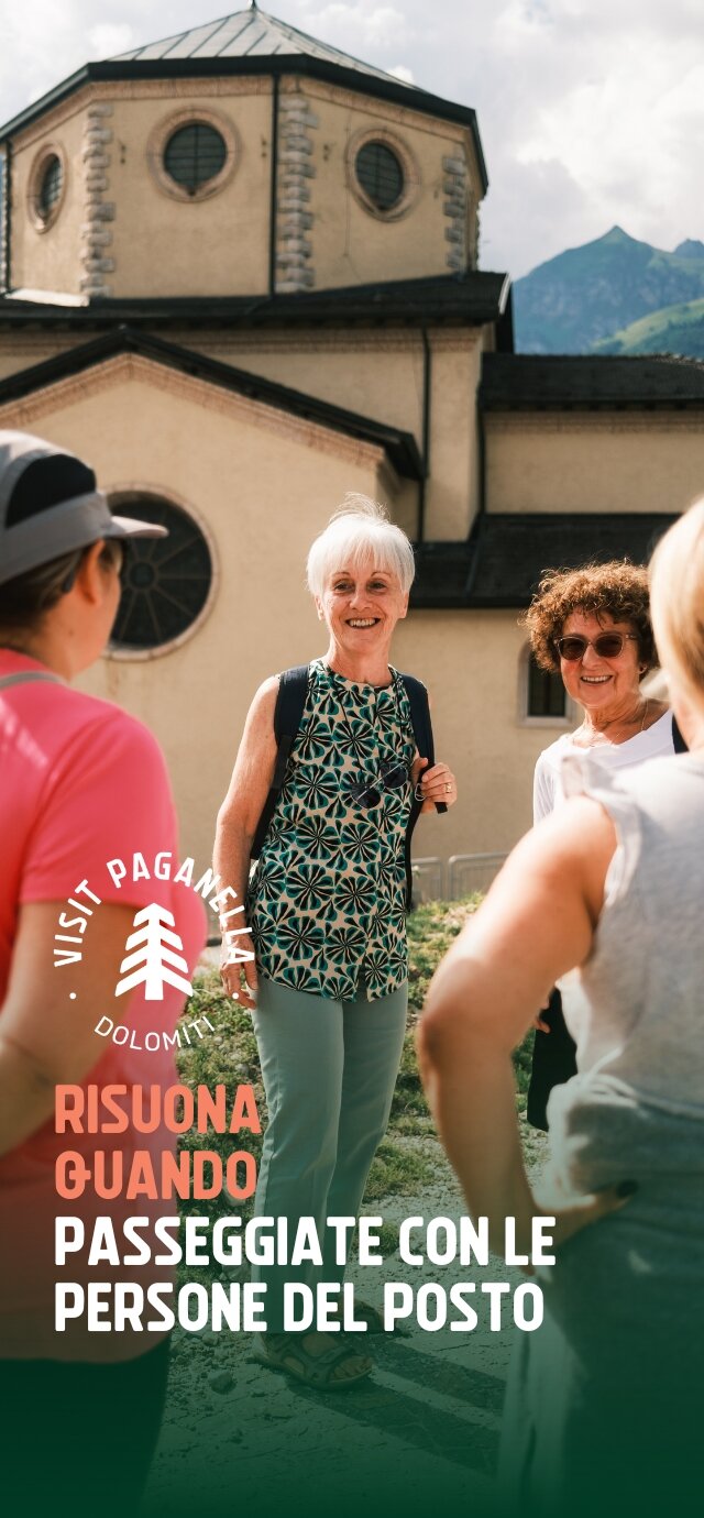 Un gruppo di persone conversa allegramente davanti a una chiesa con una grande cupola e finestre circolari. La scena è illuminata dalla luce del sole. Una donna anziana con uno zaino sorride mentre parla con altre persone. Le montagne fanno da sfondo. Il testo sulla foto recita: "Risuona quando passeggiate con le persone del posto." | © Filippo Frizzera, 2024