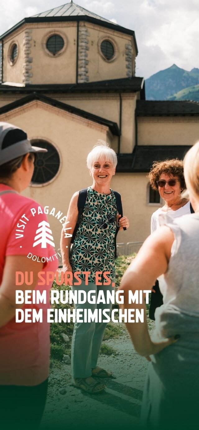 Eine Gruppe von Menschen unterhält sich fröhlich vor einer Kirche mit einer großen Kuppel und runden Fenstern. Die Szene wird vom Sonnenlicht beleuchtet. Eine ältere Frau mit einem Rucksack lächelt, während sie sich mit anderen Menschen unterhält. Die Berge bilden den Hintergrund. Der Text auf dem Foto lautet: „Du spürst es beim Rundgang mit den Einheimischen“. | © Filippo Frizzera, 2024