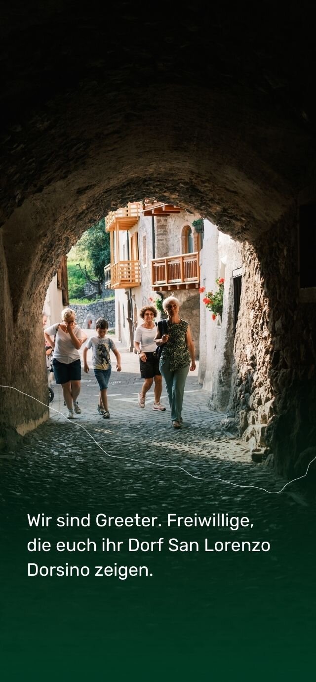 Ein Steintunnel mit einer dunklen, rauen Decke öffnet sich zu einer sonnigen Gasse. Am Ende stehen Berghäuser mit Holzbalkonen und roten Blumen in den Fenstern. Eine kleine Gruppe von Menschen geht lächelnd auf den Ausgang des Bogens zu. Eine ältere Frau im Vordergrund scheint die Gruppe anzuführen. Der Text lautet: „Wir sind Greeter. Freiwillige, die euch ihr Dorf San Lorenzo Dorsino zeigen.“ | © Filippo Frizzera, 2024