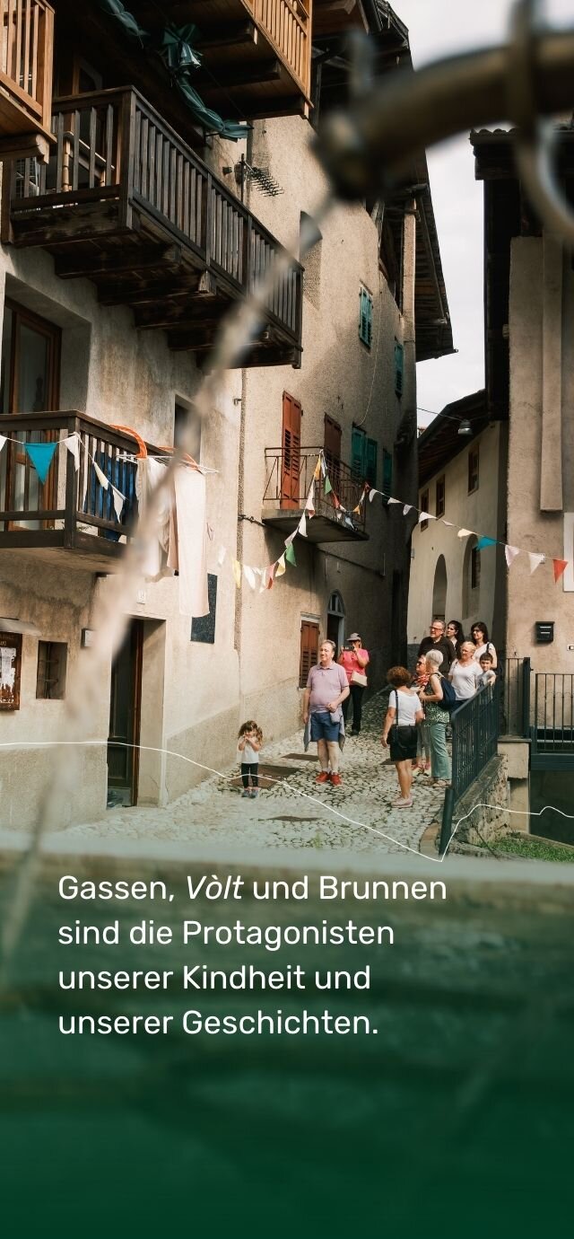 Ein Brunnen im Vordergrund gibt den Blick auf ein altes Dorf frei. Im Hintergrund: gepflasterte Gassen, Steinhäuser mit Holzbalkonen und bunten Fenstern. Fahnengirlanden durchziehen die Straße. Eine Gruppe von Menschen, darunter Erwachsene und Kinder, schlendert die Gasse entlang. Ein kleines Mädchen hält sich die Hände vor das Gesicht, als würde es spielen oder sich vor der Sonne schützen. Der Text auf dem Foto lautet: „Gassen, Vòlt und Brunnen sind die Protagonisten unserer Kindheit und unserer Geschichten“. | © Filippo Frizzera, 2024