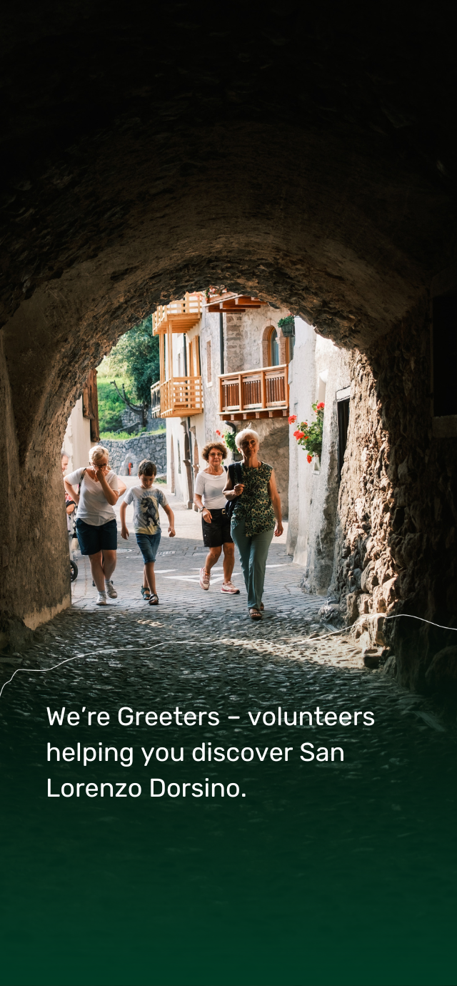 A stone tunnel with a dark, rough ceiling opens onto a sunny alley. At the end, mountain houses with wooden balconies and red flowers in the windows. A small group of people walk towards the exit of the arch, smiling. An elderly woman, in the foreground, seems to lead the group. The text reads: ‘We're Greeters - volunteers helping you discover San Lorenzo Dorsino.' | © Filippo Frizzera, 2024
