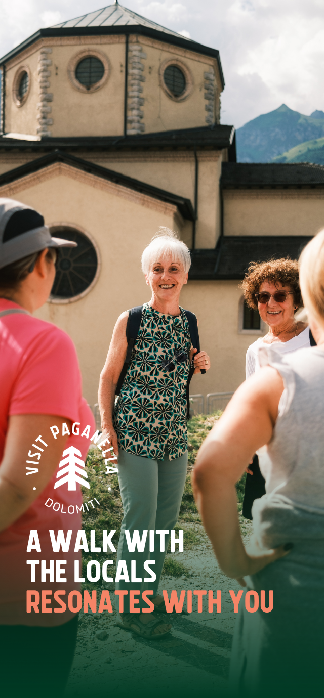 A group of people converse happily in front of a church with a large dome and circular windows. The scene is illuminated by sunlight. An elderly woman with a backpack smiles as she talks to other people. Mountains form the background. The text on the photo reads: 'A walk with the locals resonates with you' | © Filippo Frizzera, 2024