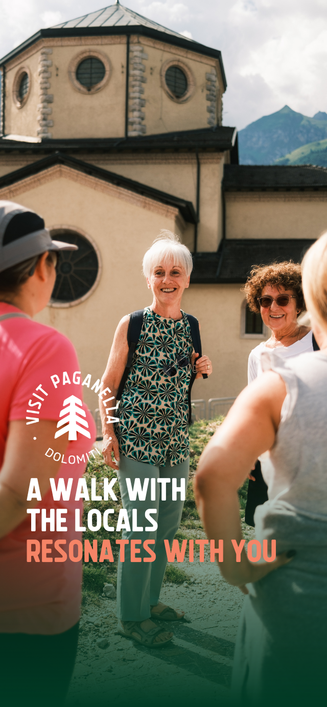 A group of people converse happily in front of a church with a large dome and circular windows. The scene is illuminated by sunlight. An elderly woman with a backpack smiles as she talks to other people. Mountains form the background. The text on the photo reads: 'A walk with the locals resonates with you' | © Filippo Frizzera, 2024