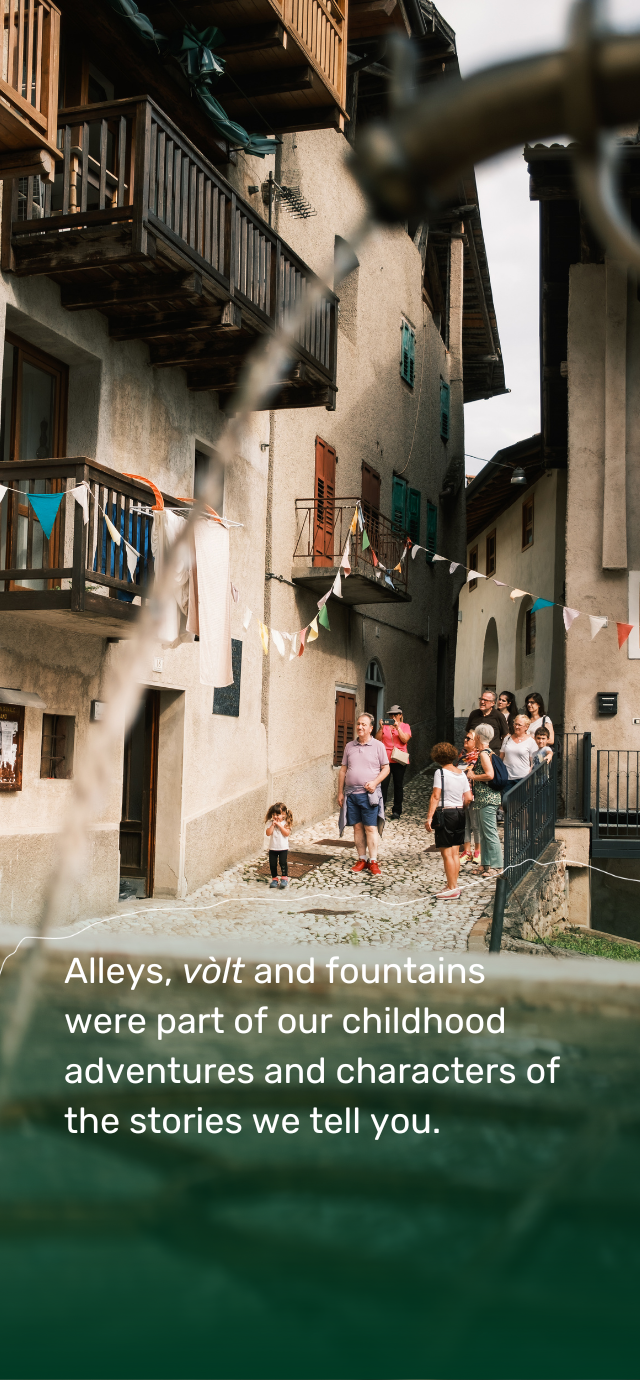 A fountain in the foreground frames a glimpse of an ancient village. In the background, cobbled alleys, stone houses with wooden balconies and colourful windows. Festoons of flags cross the street. A group of people, including adults and children, stroll down the alley. A little girl with her hands over her face seems to be playing or protecting herself from the sun. The text on the photo reads: ‘Alleys, vòlt and fountains were part of our childhood adventures and characters of the stories we tell you’.  Translated with DeepL.com (free version) | © Filippo Frizzera, 2024